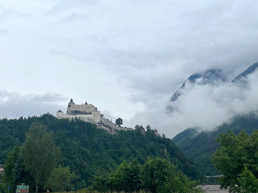 Die Burg Hohenwerfen hoch auf einem grün bewaldeten Berg, Wolken hängen im Tal