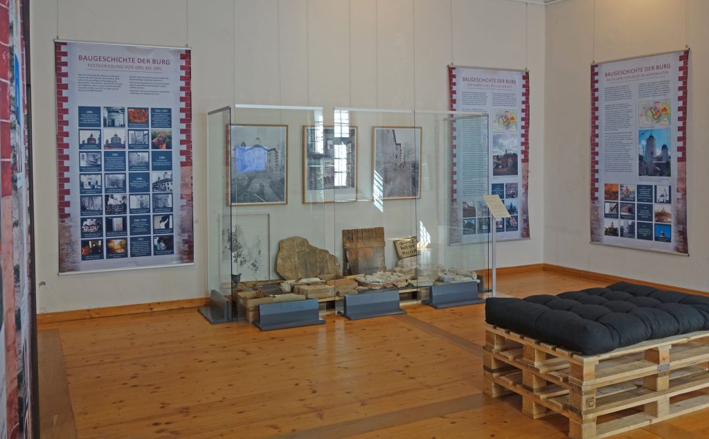 Ausstellungsraum mit Stoffbannern an den Wänden, Baustellenfunden und einer Bank aus Holzpaletten.