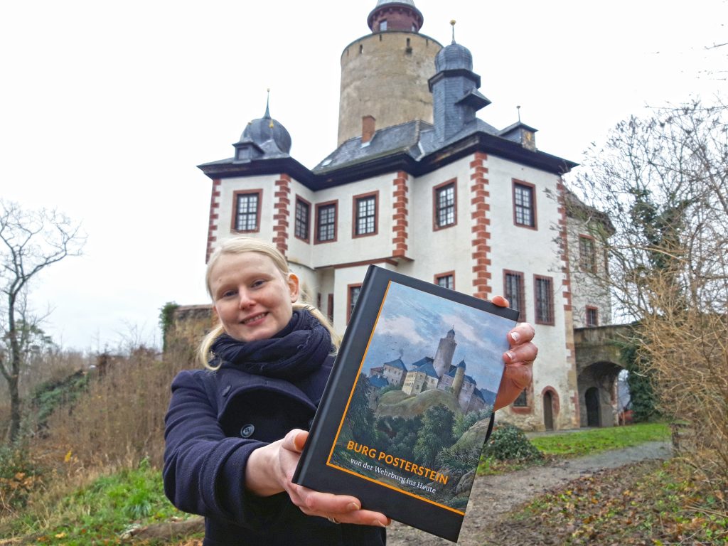 Eine junge Frau steht vor der Burg Posterstein und hält ein Buch in die Kamera