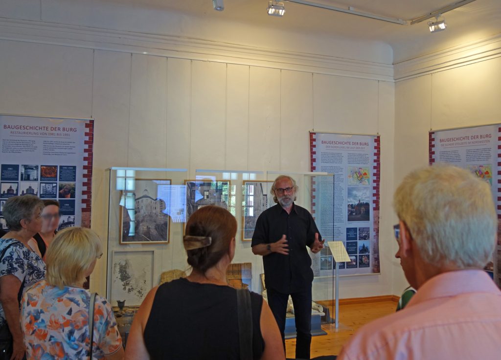 Im Vordergrund Publikum von hinten, vor den Ausstellungsbannern der Museumsdirektor bei einer Führung.