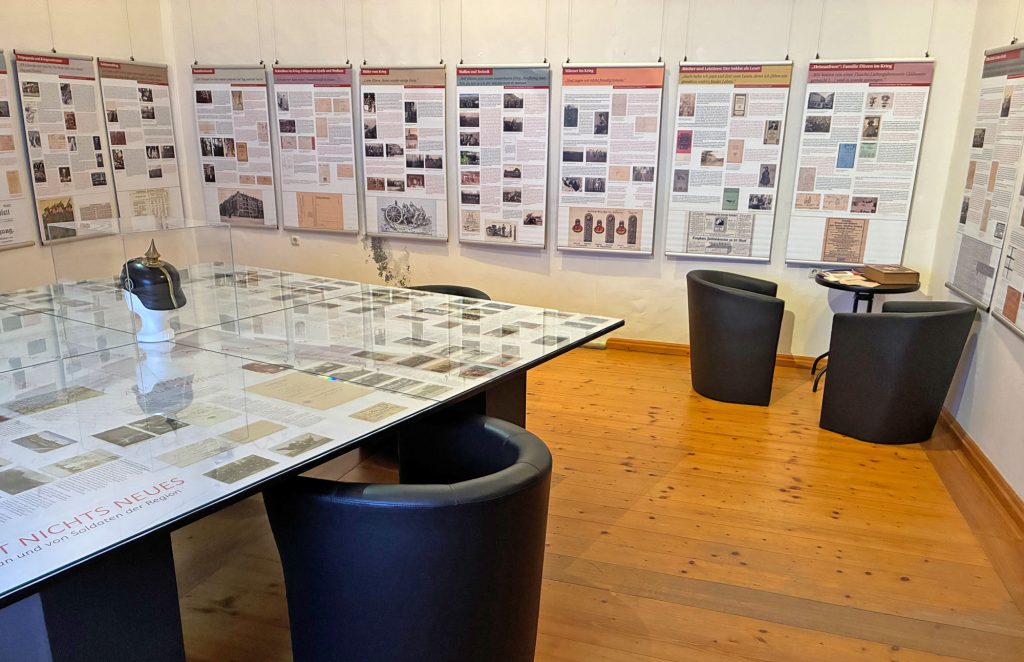 Im Vordergrund ein Tisch mit Sesseln, im Hintergrund 11 Textbanner an der Wand